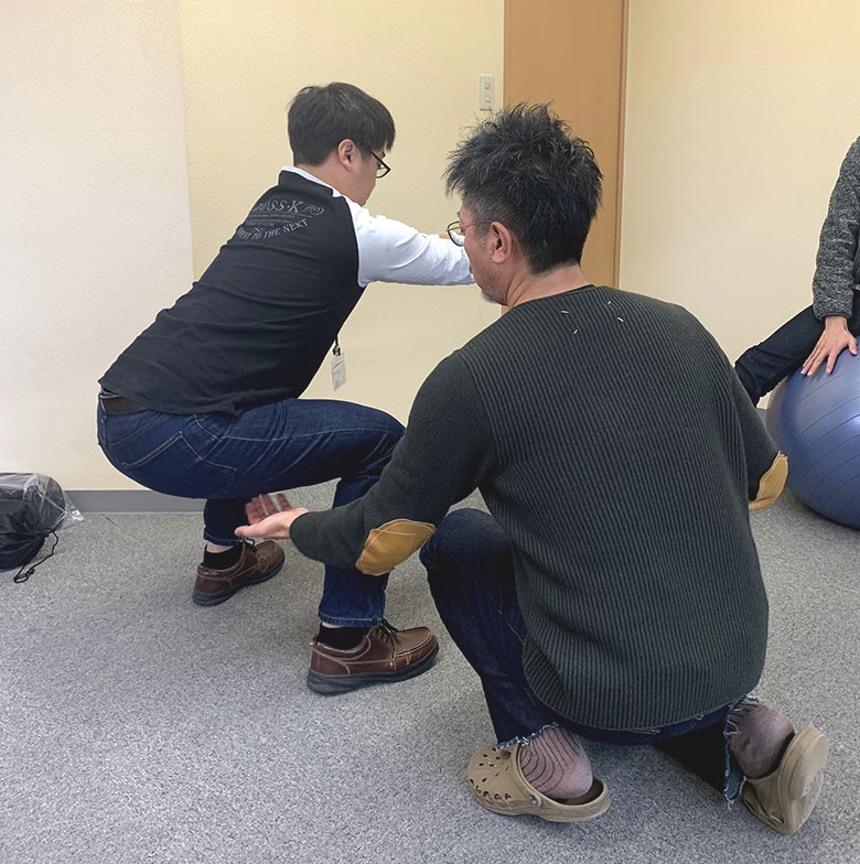 スクワットをする男性社員と、サポートする代表高村